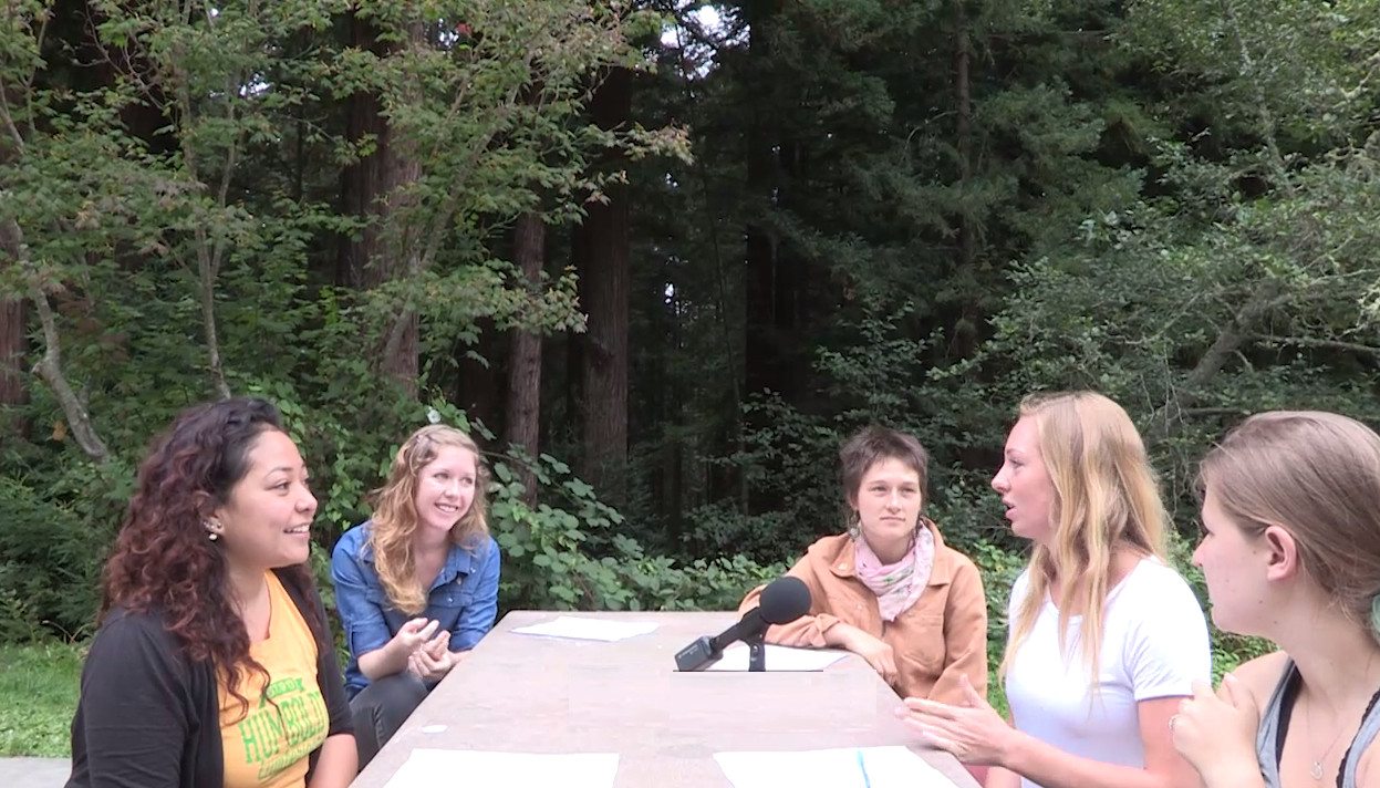 Five people sitting around a square table with a microphone talking