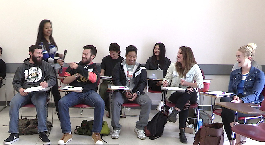 Person standing handing a microphone to another person in a classroom with several people sitting nearby at desks