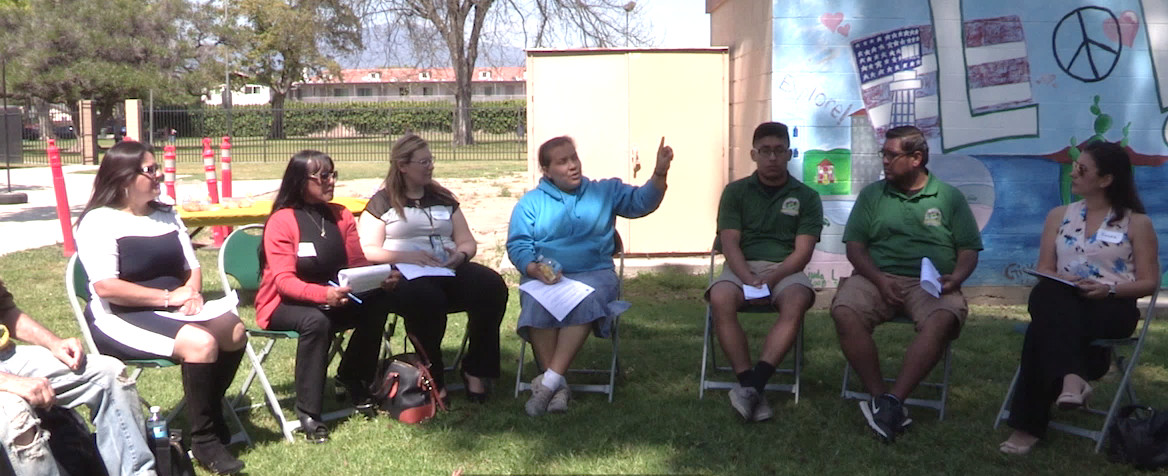 A group of people sitting on folding chairs outside looking at one person talking