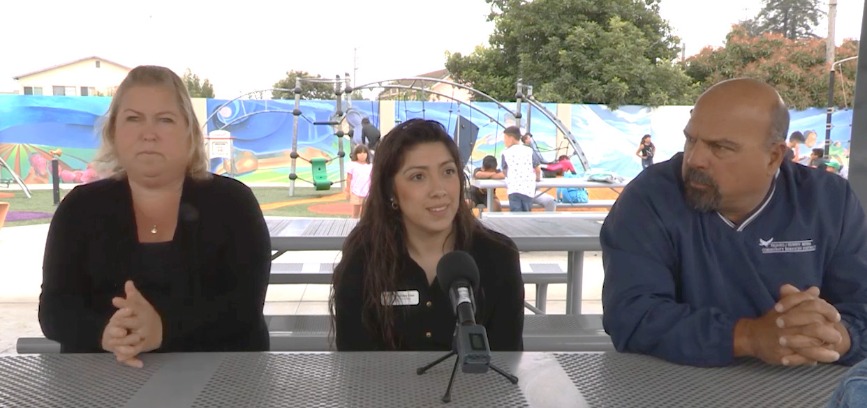 Person talking into a microphone next to two other people sitting on one side of a metal picnic table