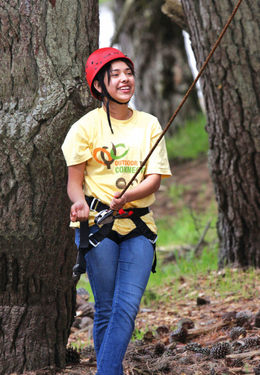A smiling woman in the forest wearing a red helmet and belaying a climber