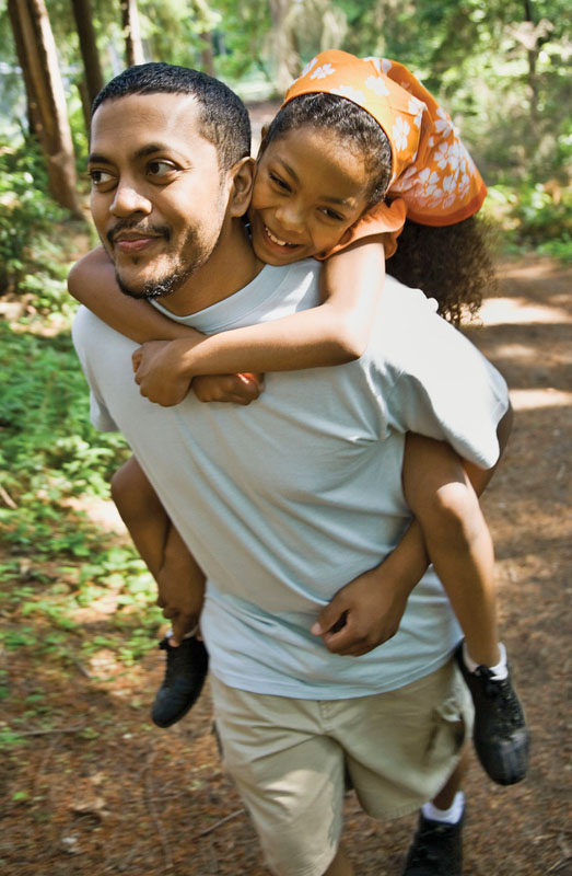 A father gives a piggy back ride to his daughter while hiking on a trail