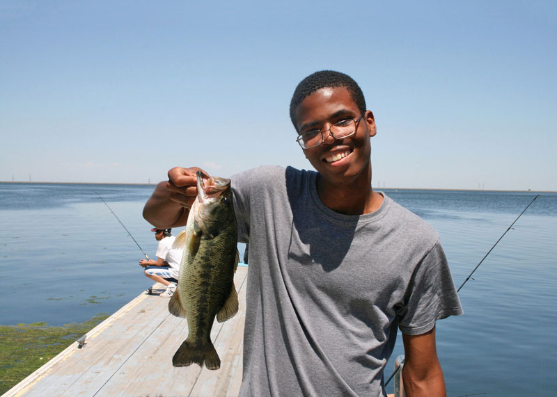 A teenager with glasses holding a bass fish he caught in the bay from a wooden dock