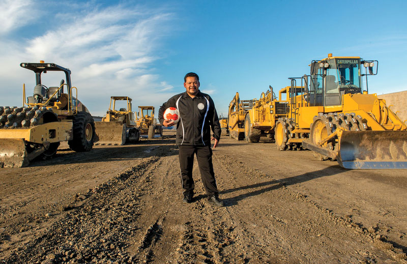A man holds a soccer ball flanked by heavy equipment used to create a new park and soccer pitch