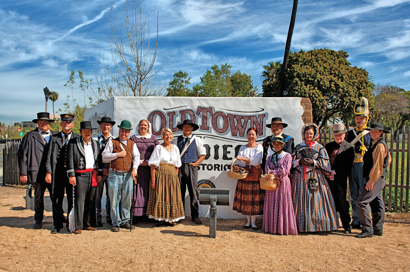 A group of men and women wear reenactment clothing and stand in front of an Old Town San Diego sign