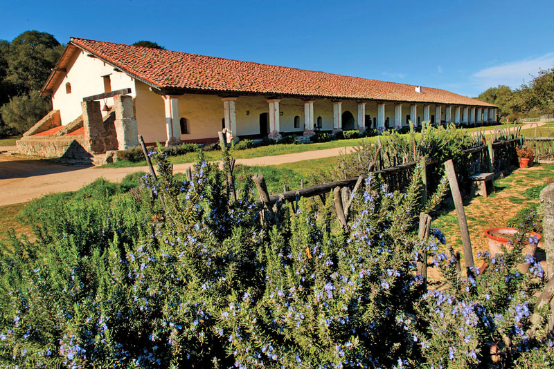 A long cream colored building with red clay tile roof and a garden in the foreground