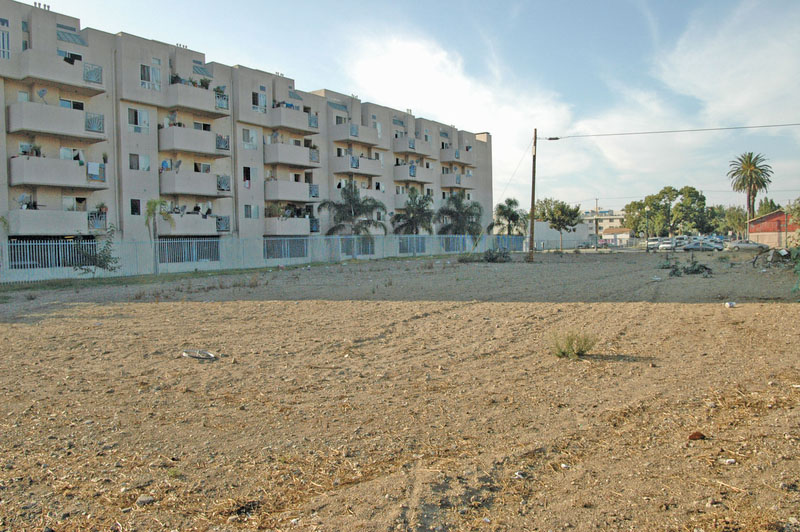 A barren vacant lot with a fence around it with an apartment complex in the background