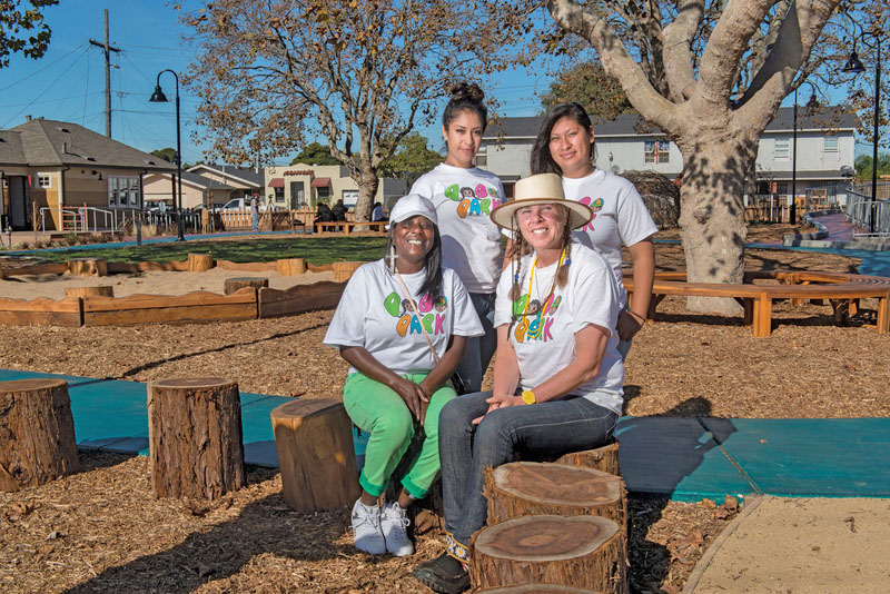 Four women, two sitting on stumps, in a new park withn shade trees in the background