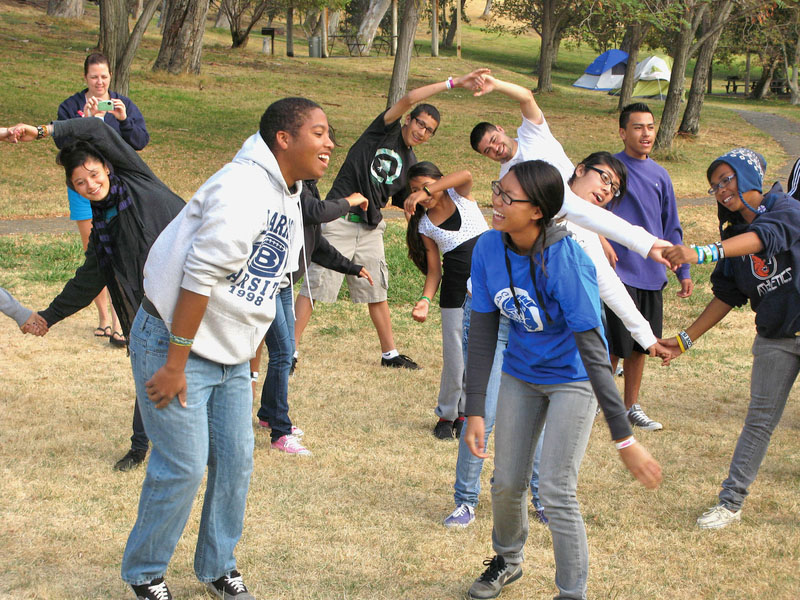 A group of teenagers dancing in a field with trees and tents in the background