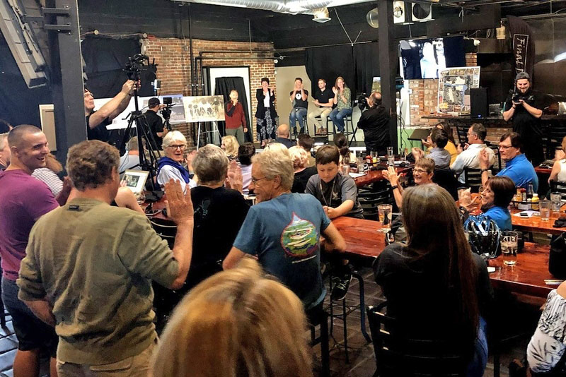 Group of people sitting, standing and talking with a group of five presenters with printed boards