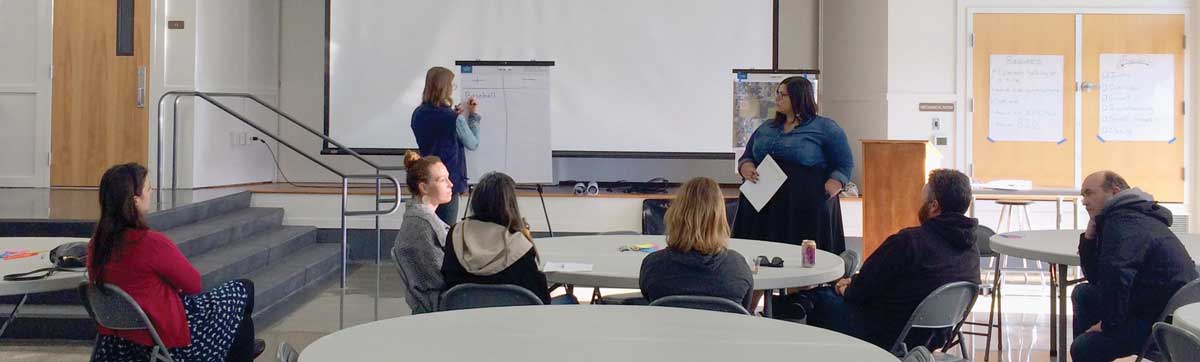 Woman writes on a large paper as a group of seven people sitting look on.