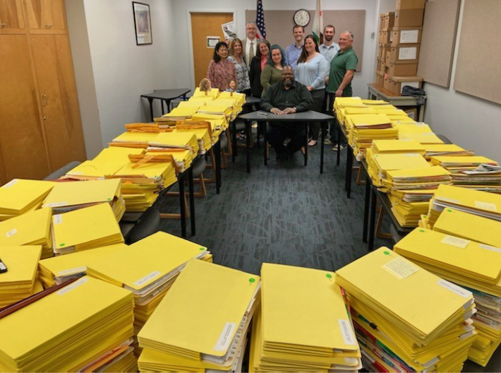 10 individuals posing next to long tables covered with towers of yellow folders.