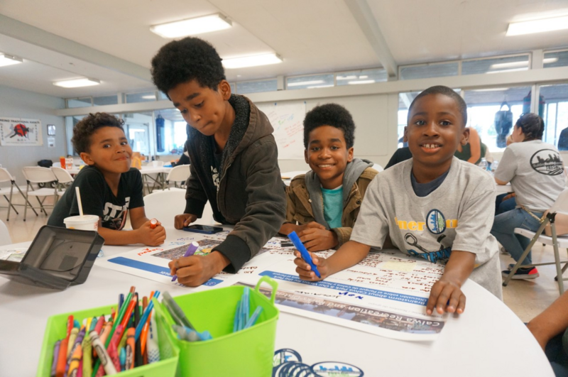 Young boys sitting on a round table writing on a large print of a park project area.