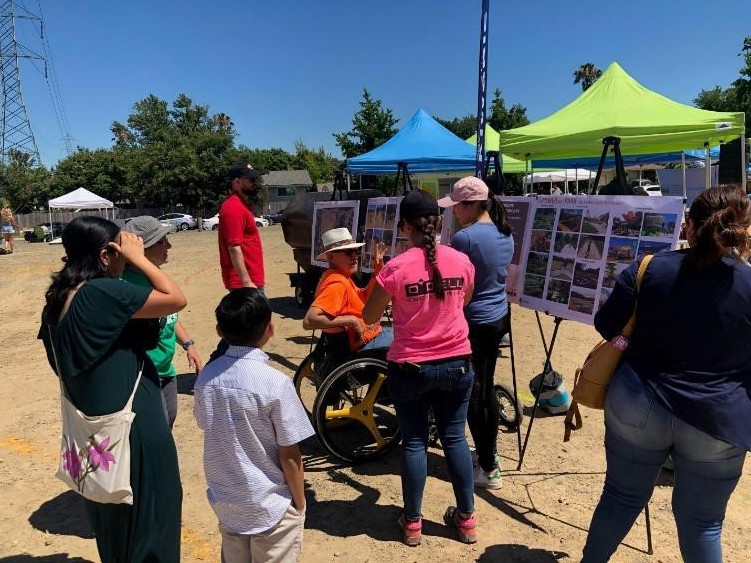 Man on wheelchair points to standing board with ideas for park features as he talks to two people.