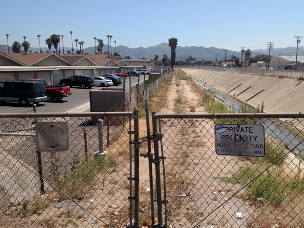 Narrow empty lot behind chainlink fence next to a river canal.