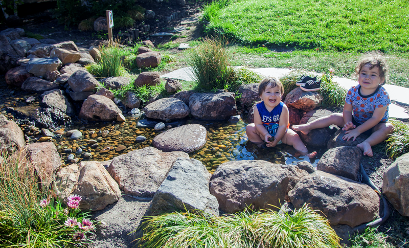 Two toddlers smile and sit on rocks near a pond.
