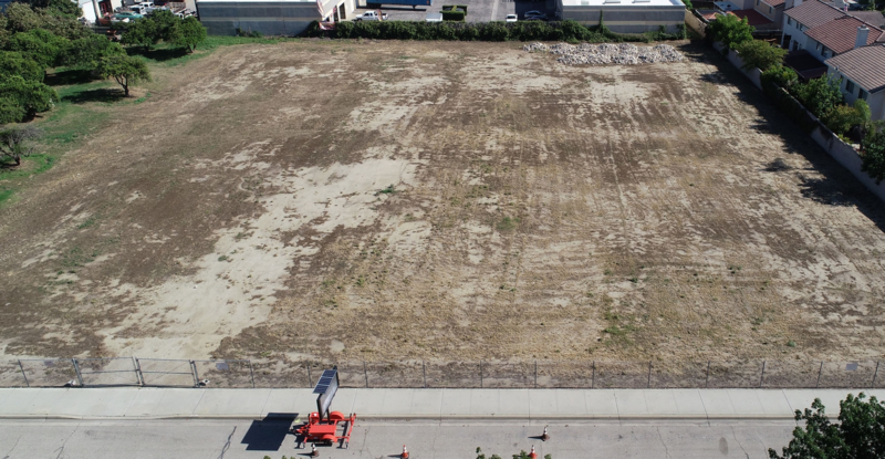 Aerial view of a vacant dirt lot enclosed by chainlink fence and surrounded by trees.