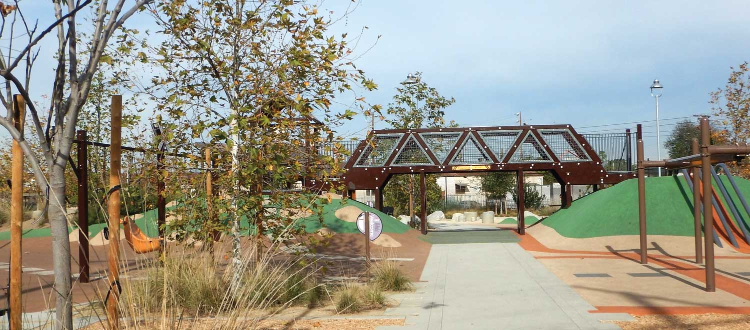 Cement walkway surrounded by trees, rubber-covered play knolls and small overhanging bridge.