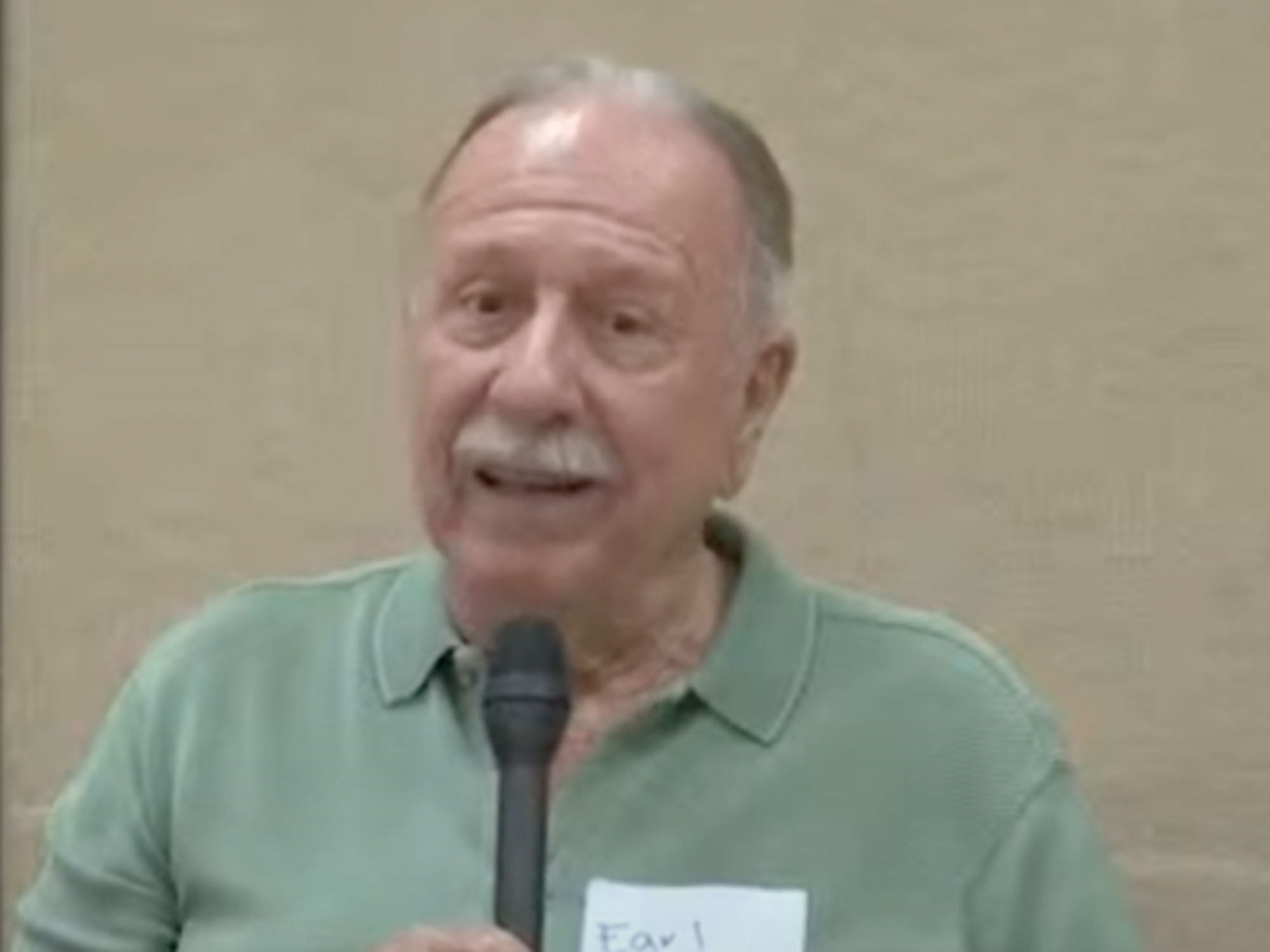 A man with a mustache wearing a green collared shirt speaking into a microphone