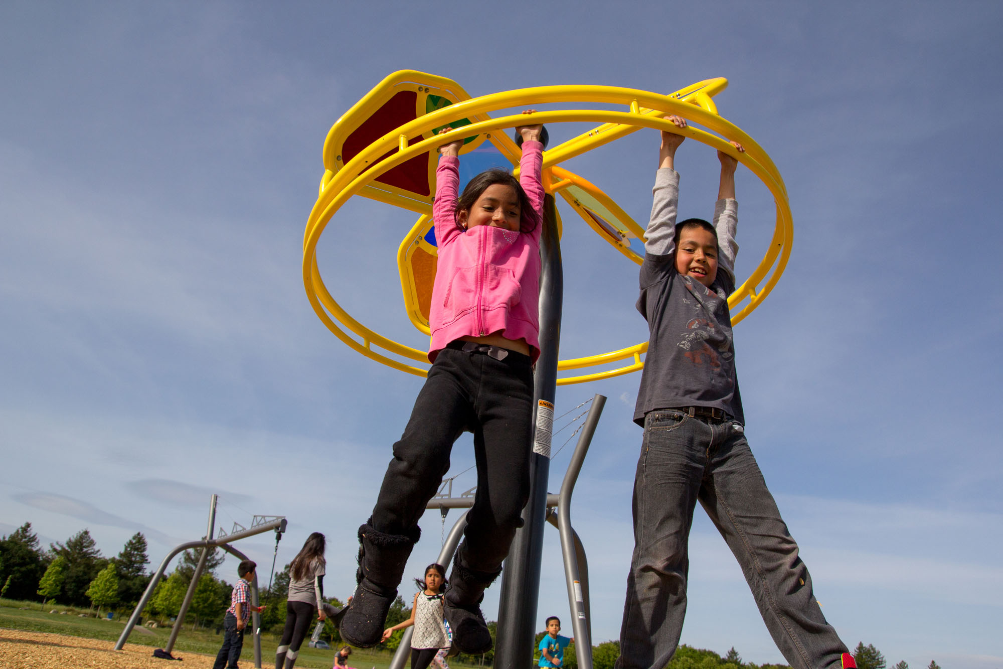 Two children hanging by their hands from a yellow spinner playground component