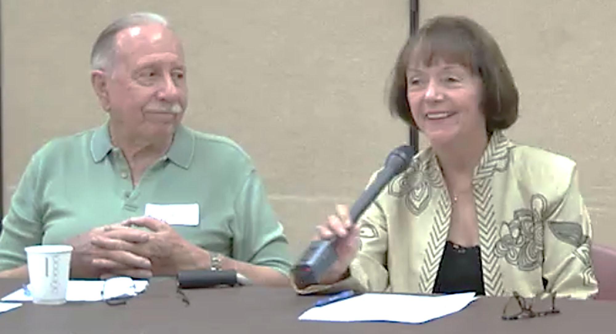 A woman smiling and speaking into a microphone during a meeting with a man seated next to her