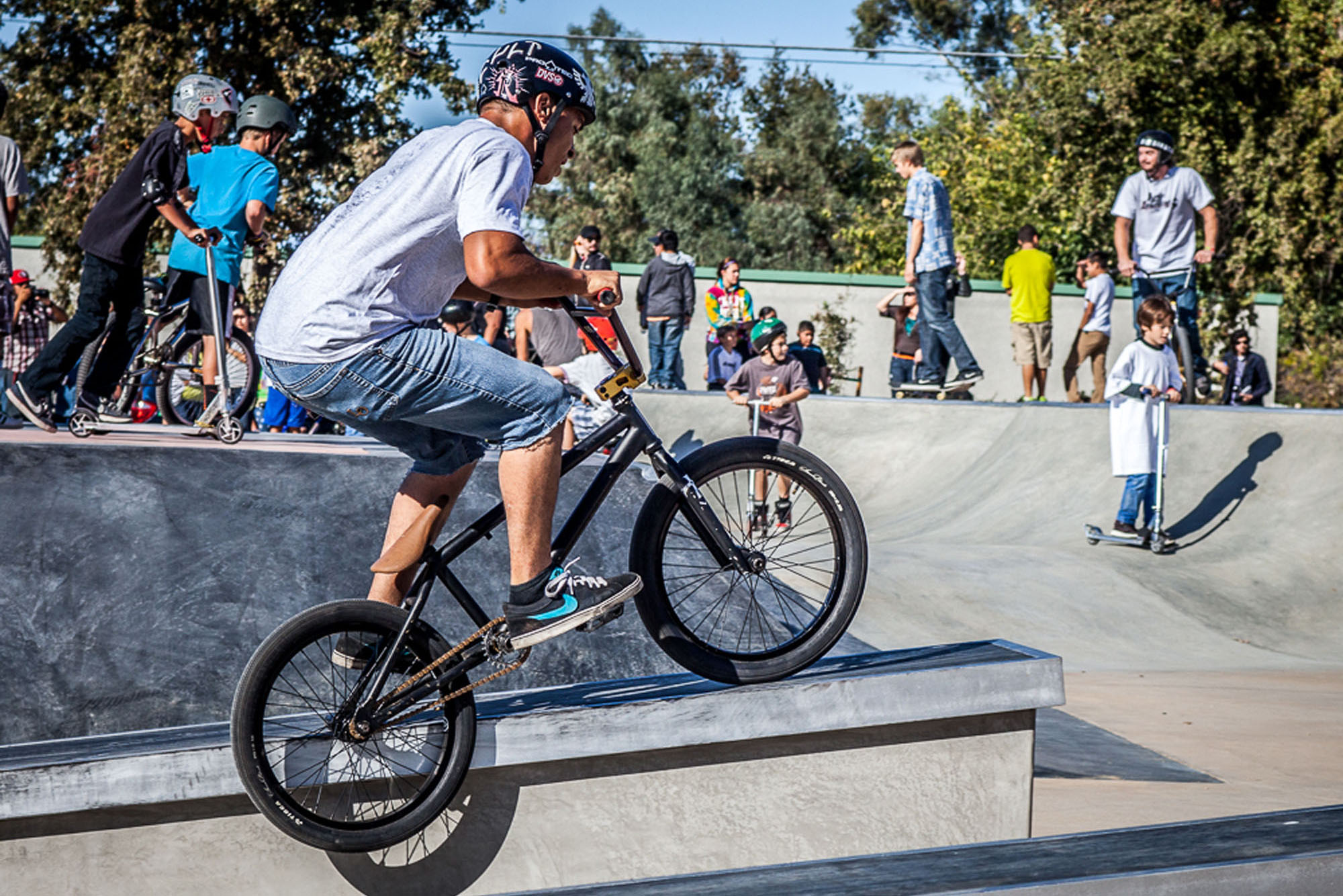 A man rides his BMX bike in a skatepark with people on scooters, bikes, and skateboards behind him