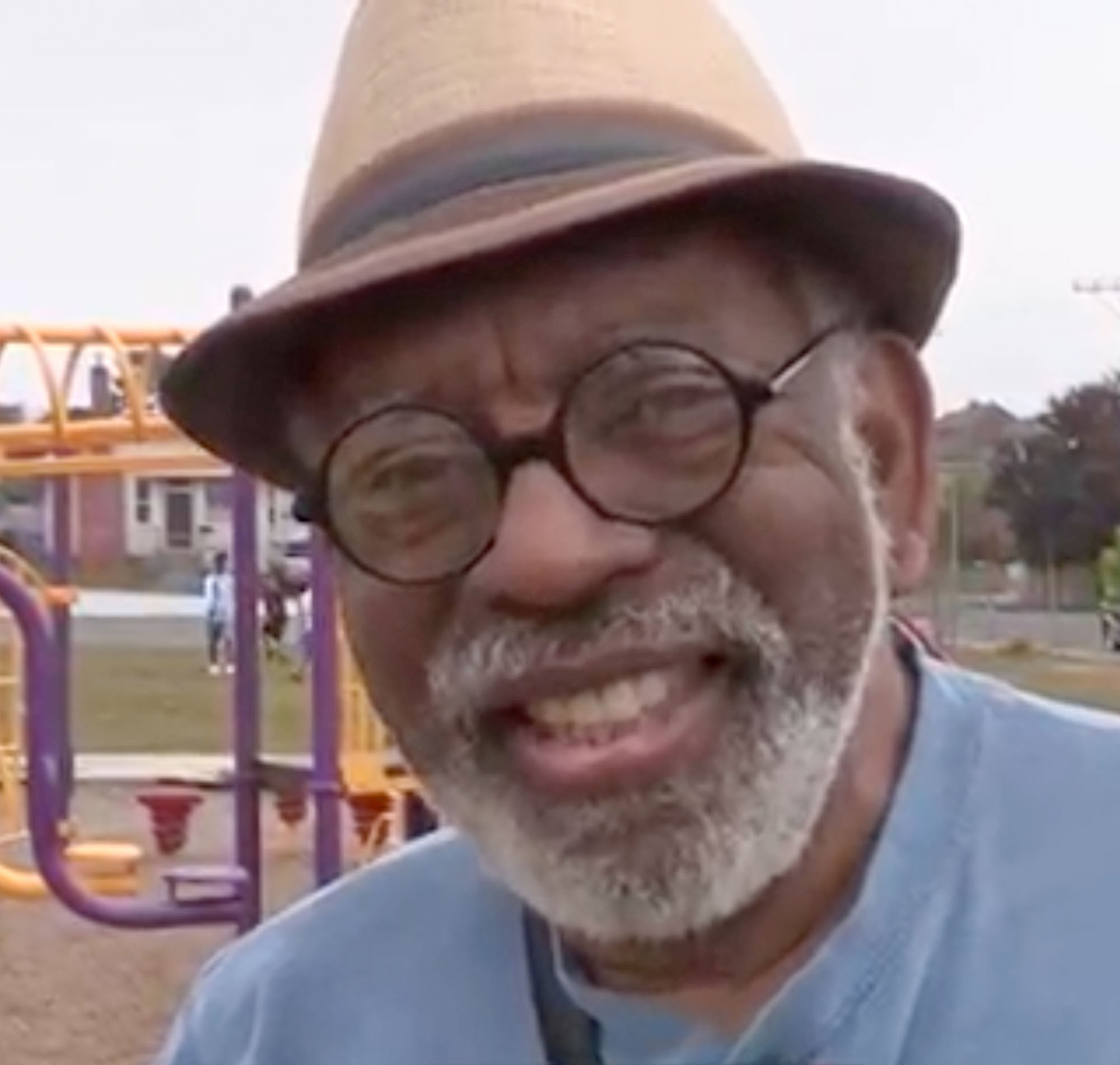A man with round glasses and a fedora smiling, standing in a playground