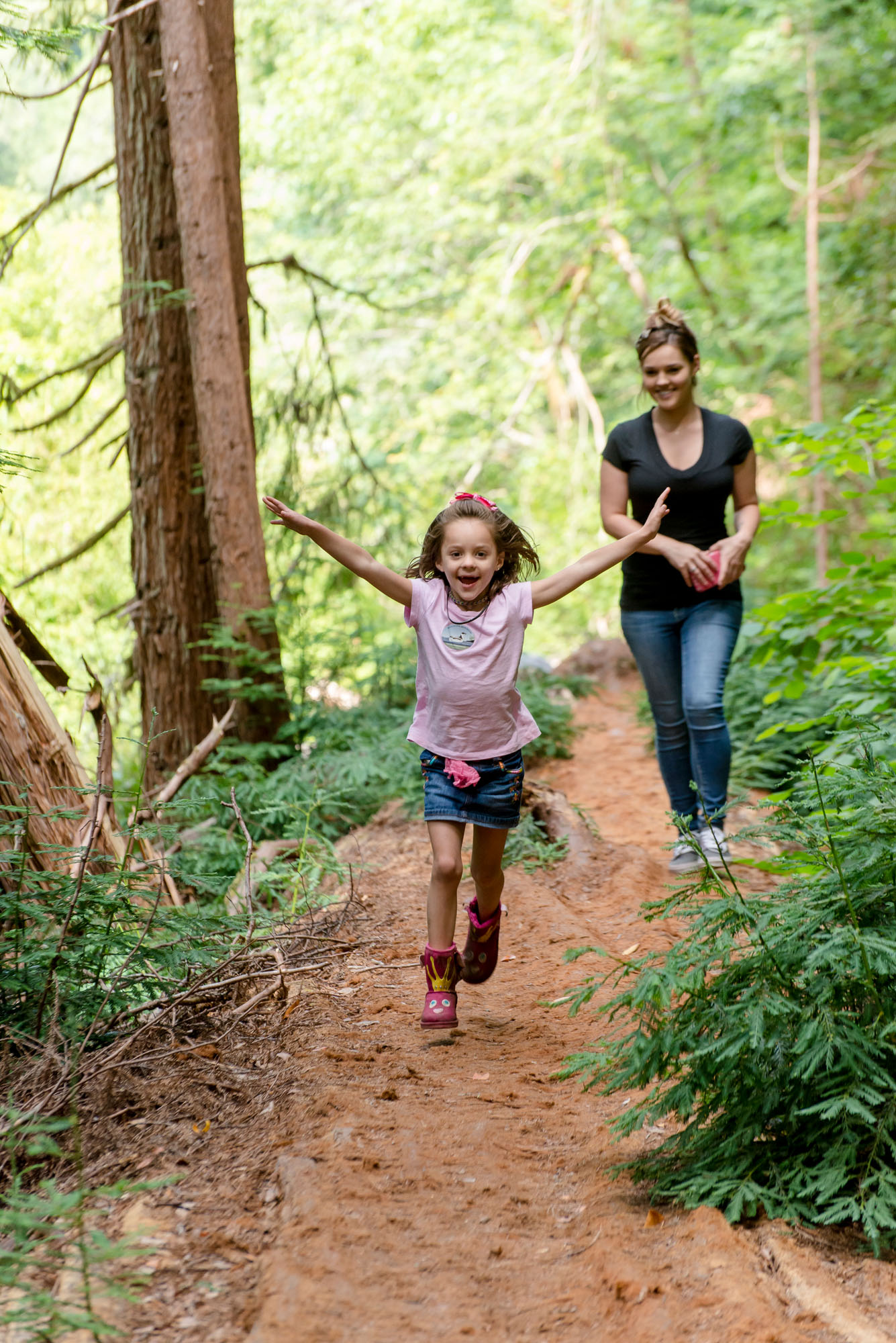 A mother and daughter hike along a dirt trail in a sun-filled forest