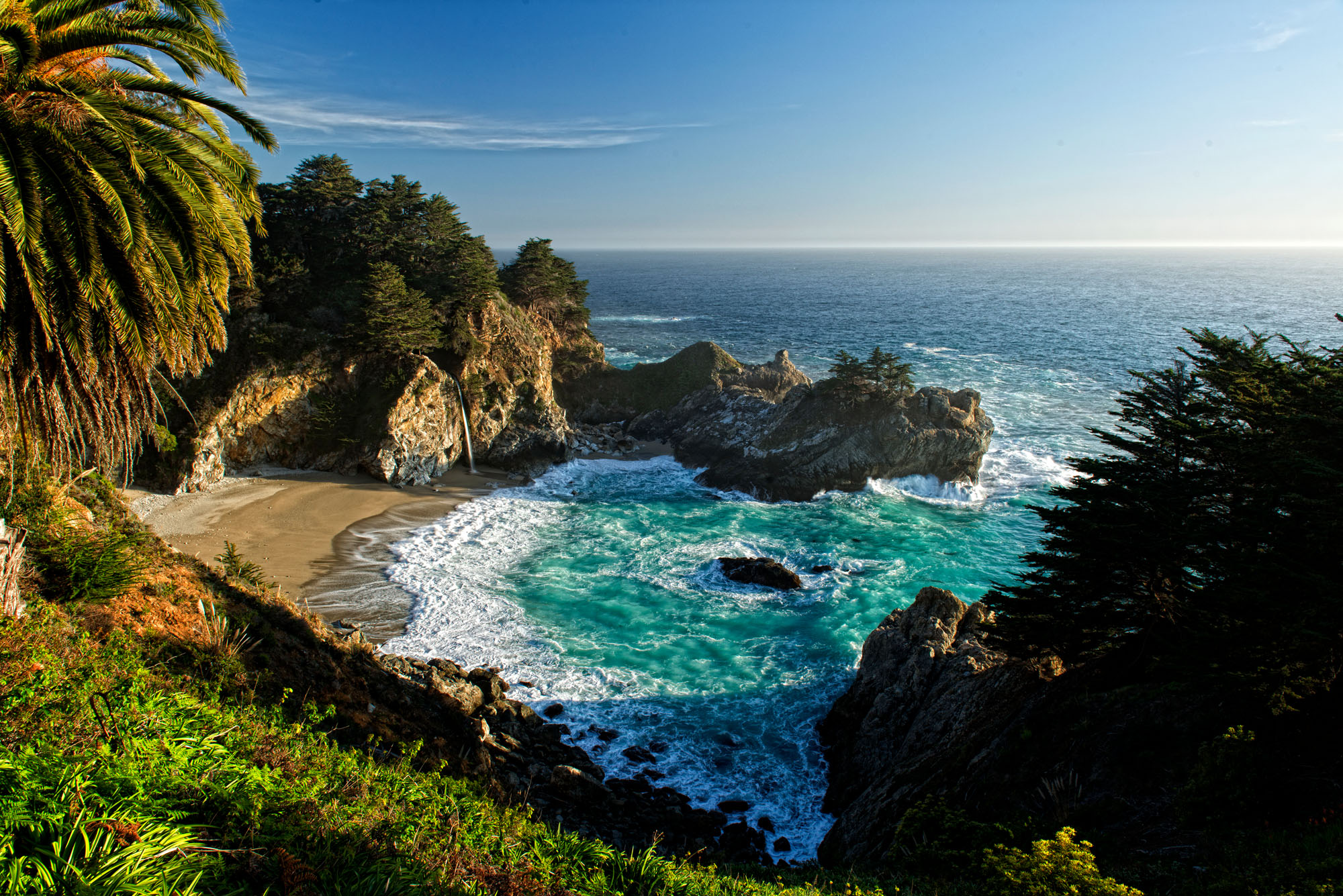 A narrow waterfall drops off of a cliff down to the sandy beach of a Pacific Ocean cove