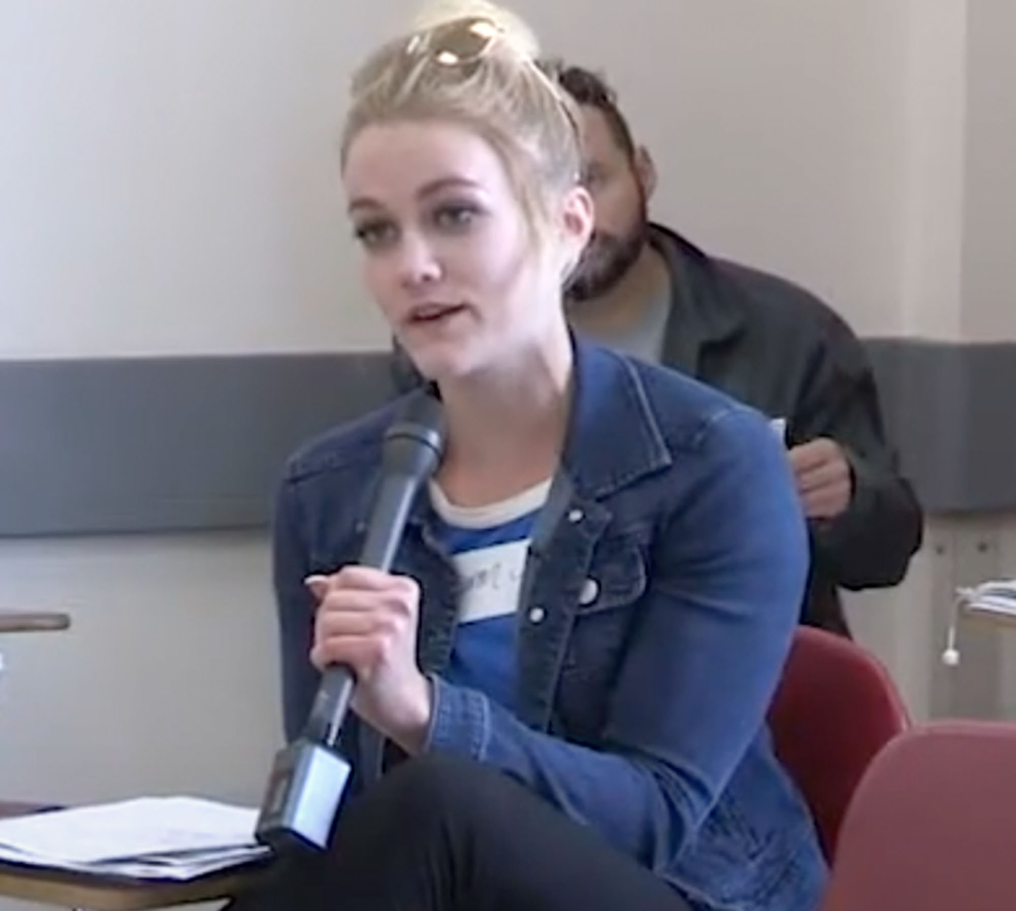 A woman with sunglasses on her head and a jean jacket speaking into a microphone in a classroom