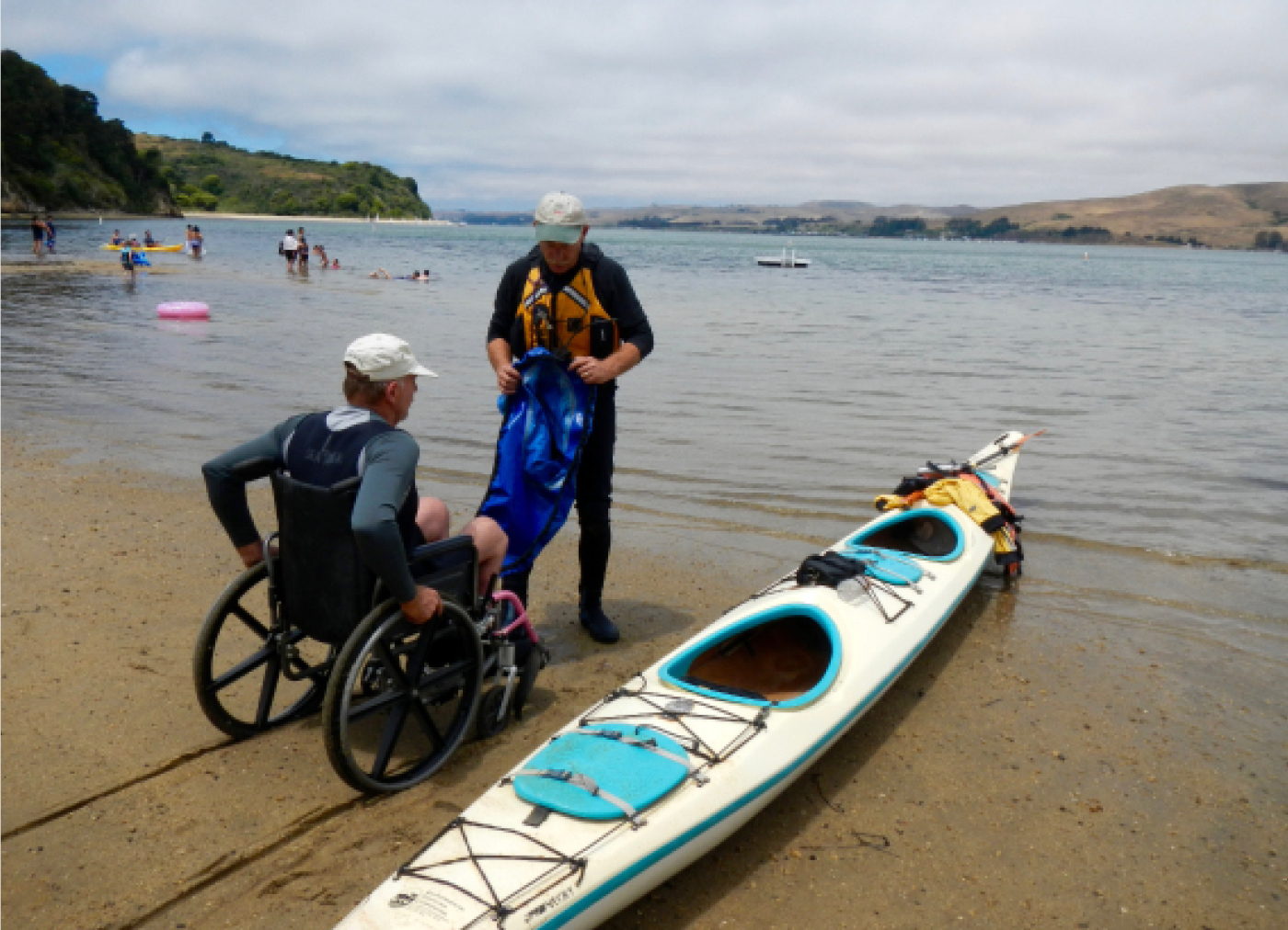 A man in a wheelchair on a sandy lake shore prepares to get into a tandem kayak with a friend