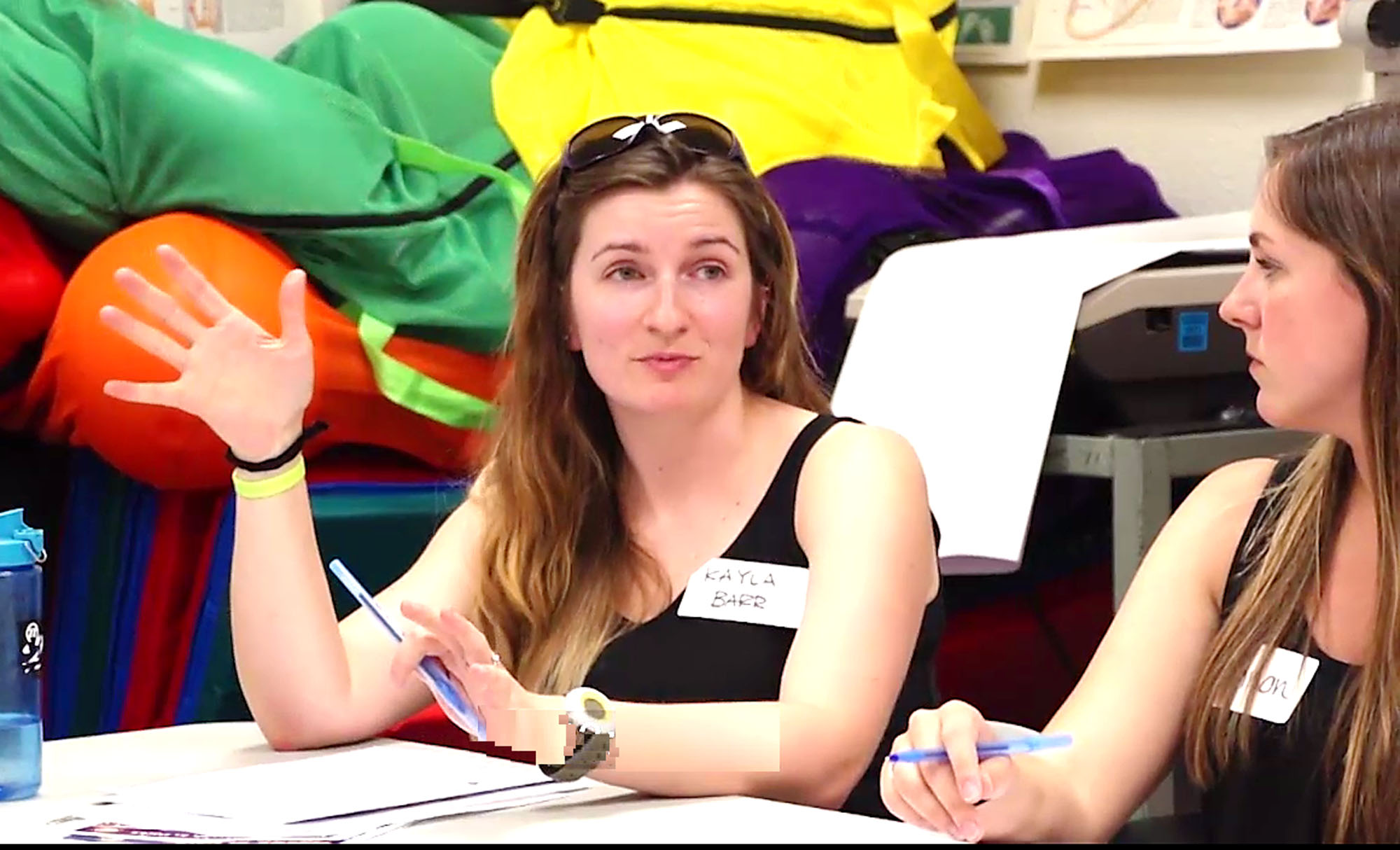 A woman with sunglasses on her head with her right hand up and a pen in her left hand in a meeting