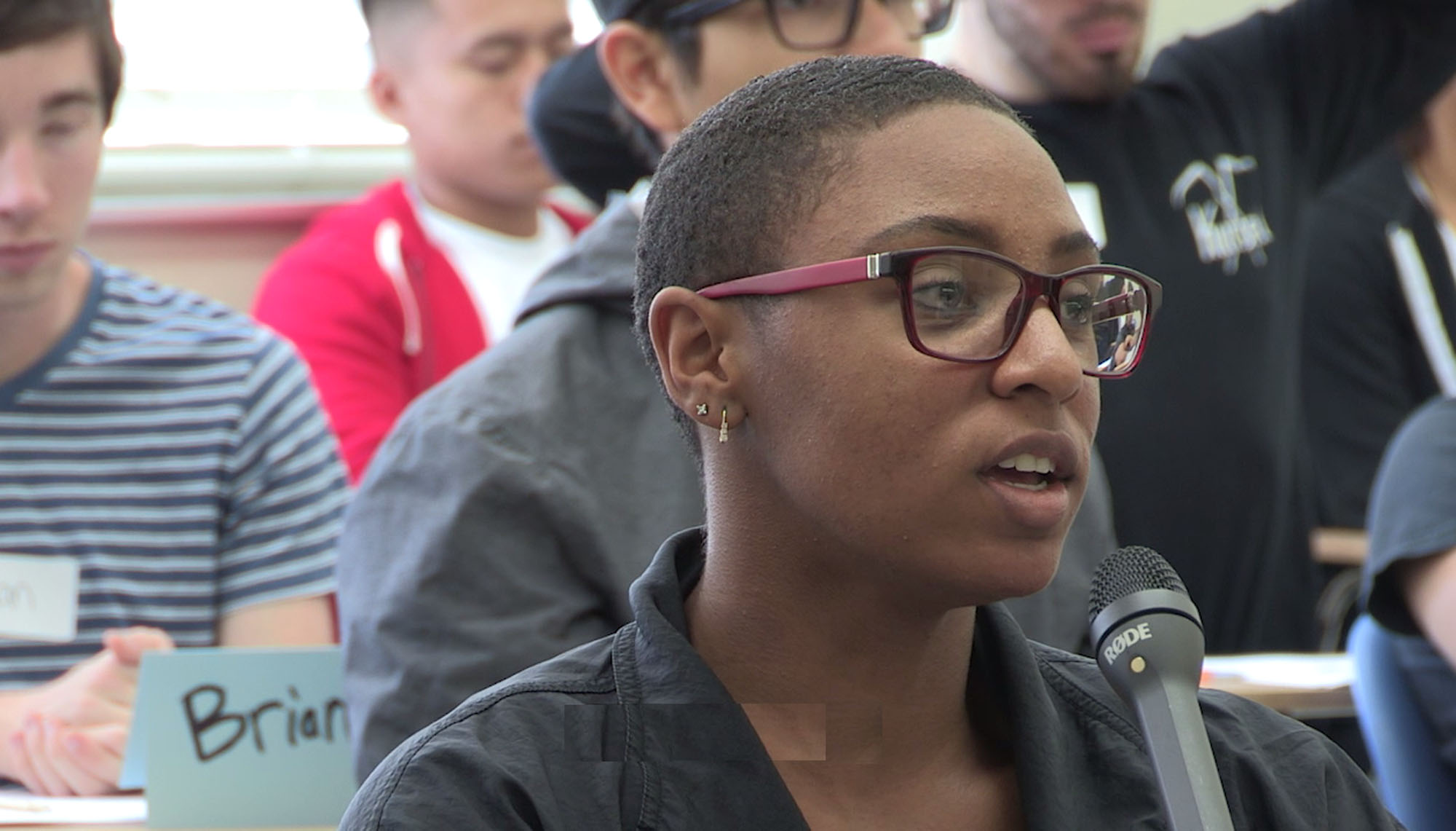 A woman with glasses speaking into a microphone with a group of people seated behind her