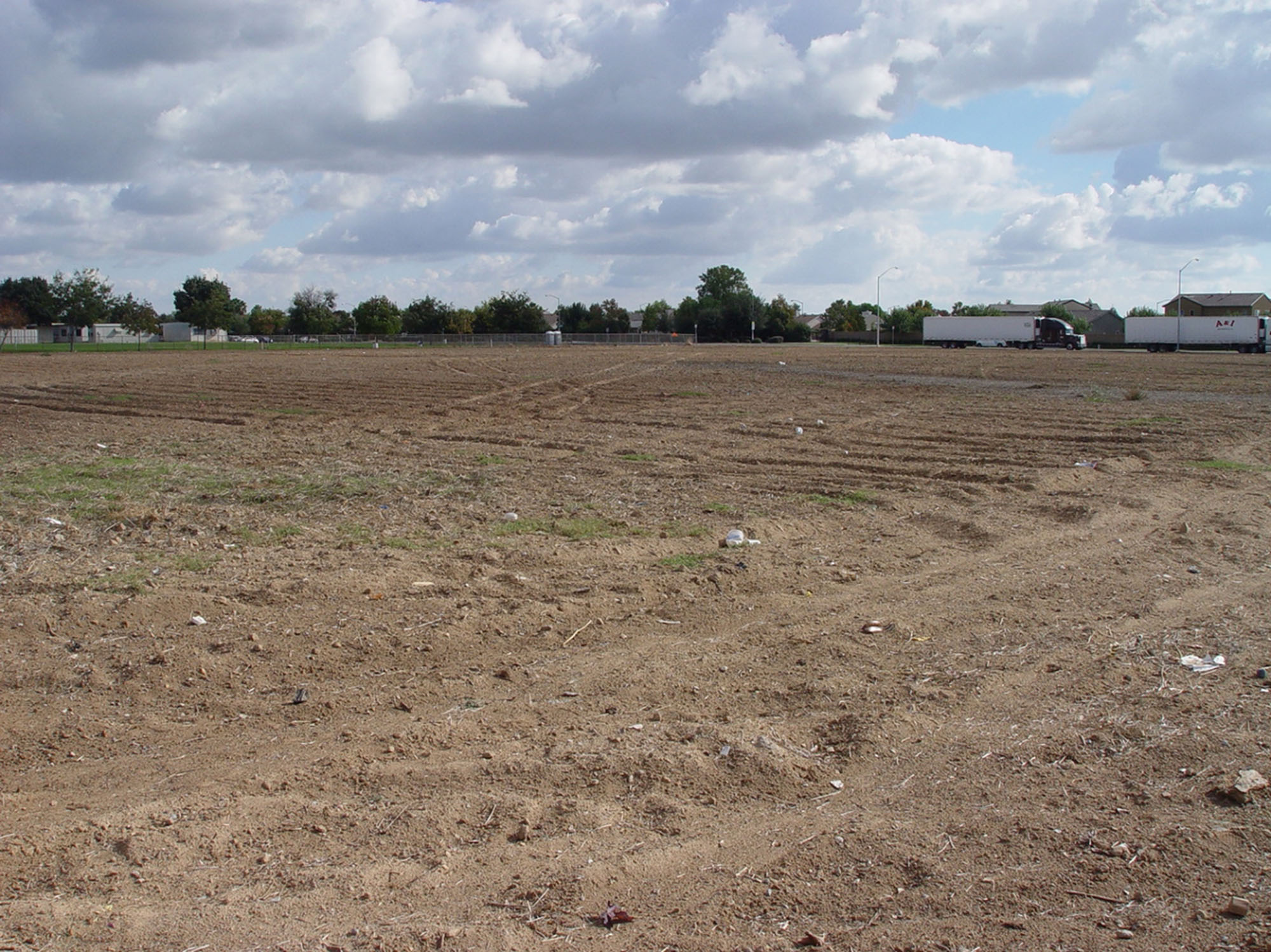 A large vacant lot, mostly dirt, patches of grass, and distributed pieces of garbage