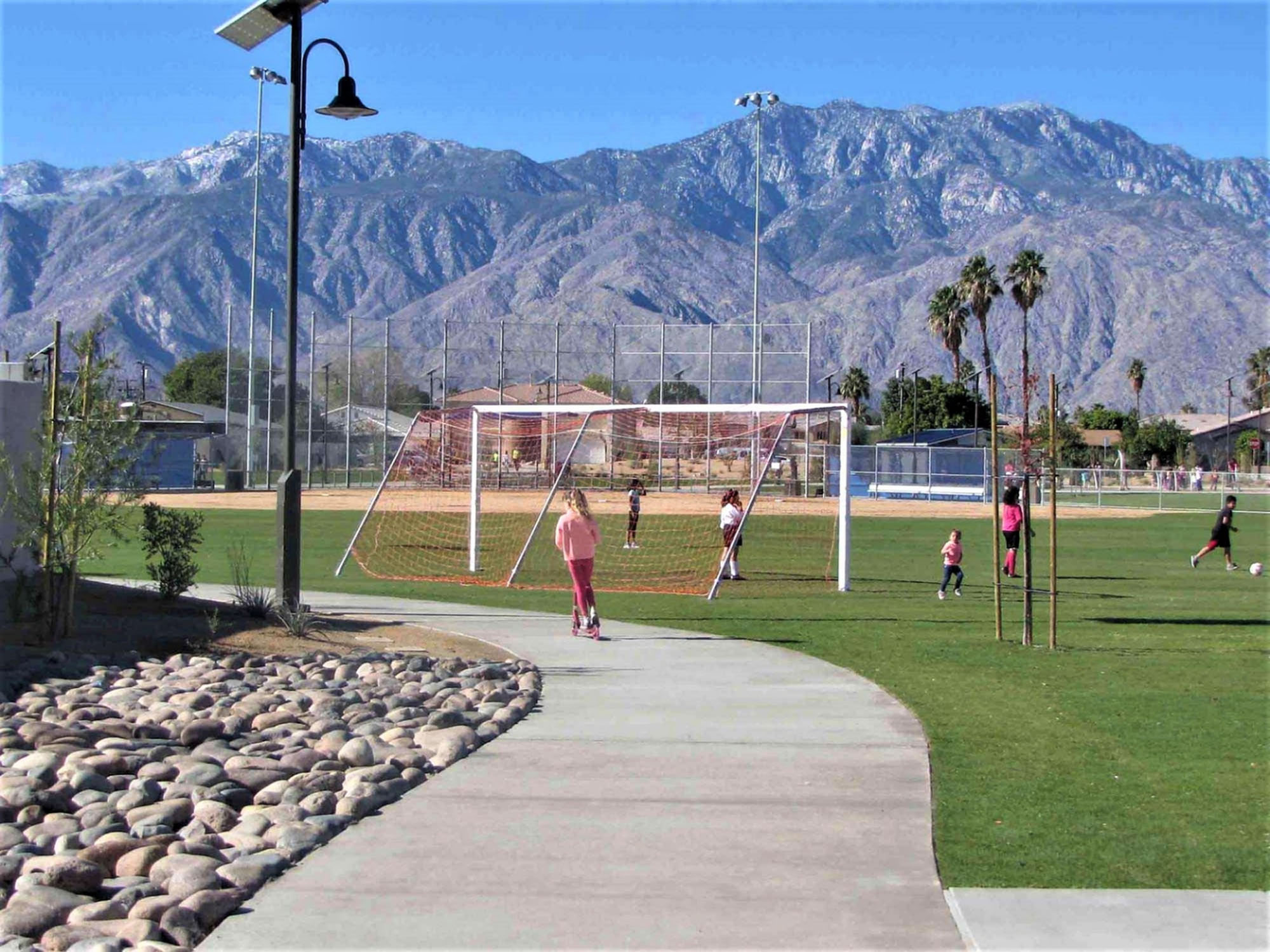 A girl on a scooter on a sidewalk in front of children playing soccer, mountains in the background