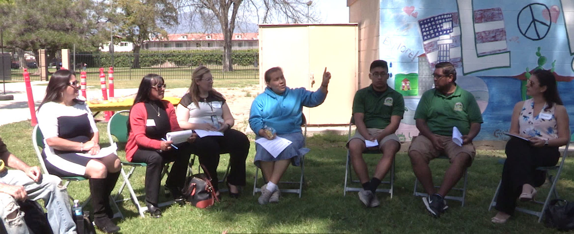 A group of people seated outside in a semicircle of chairs discussing plans for new parks