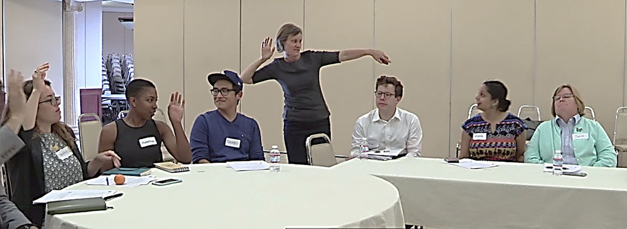 A group of people seated in a conference room with hands raised with a woman facilitating a meeting