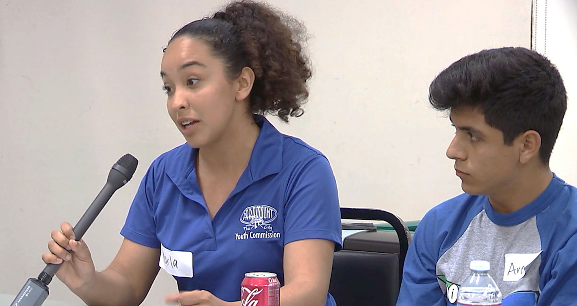 A teenage member of a youth commission speaking into a microphone with another teenager listening