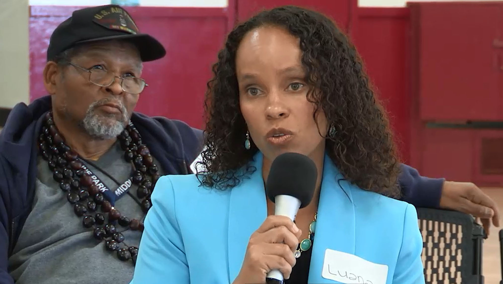 A woman with speaking into a microphone with a man seated behind her listening