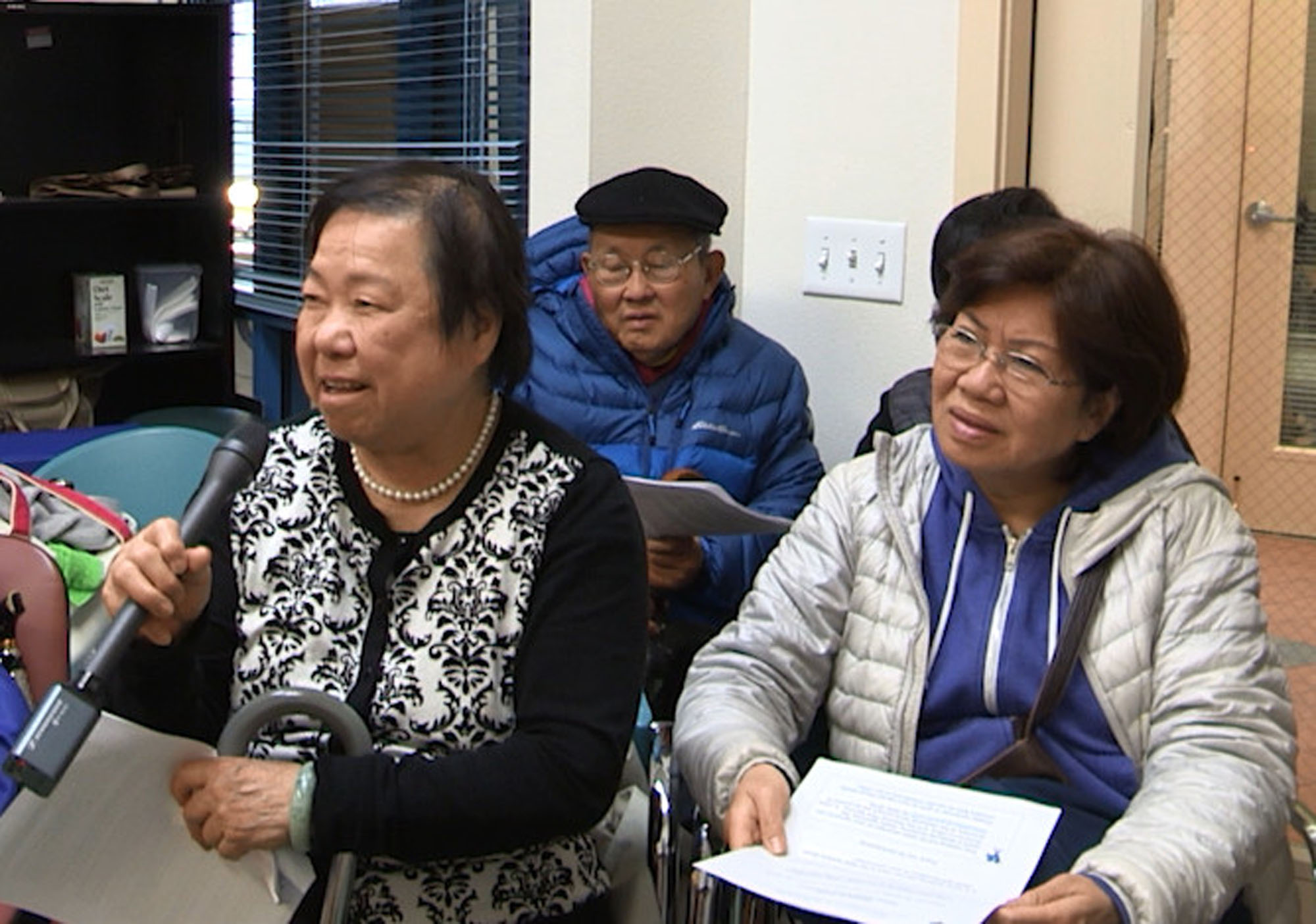 A woman with a cane speaking into a microphone with a few people sitting around her listening