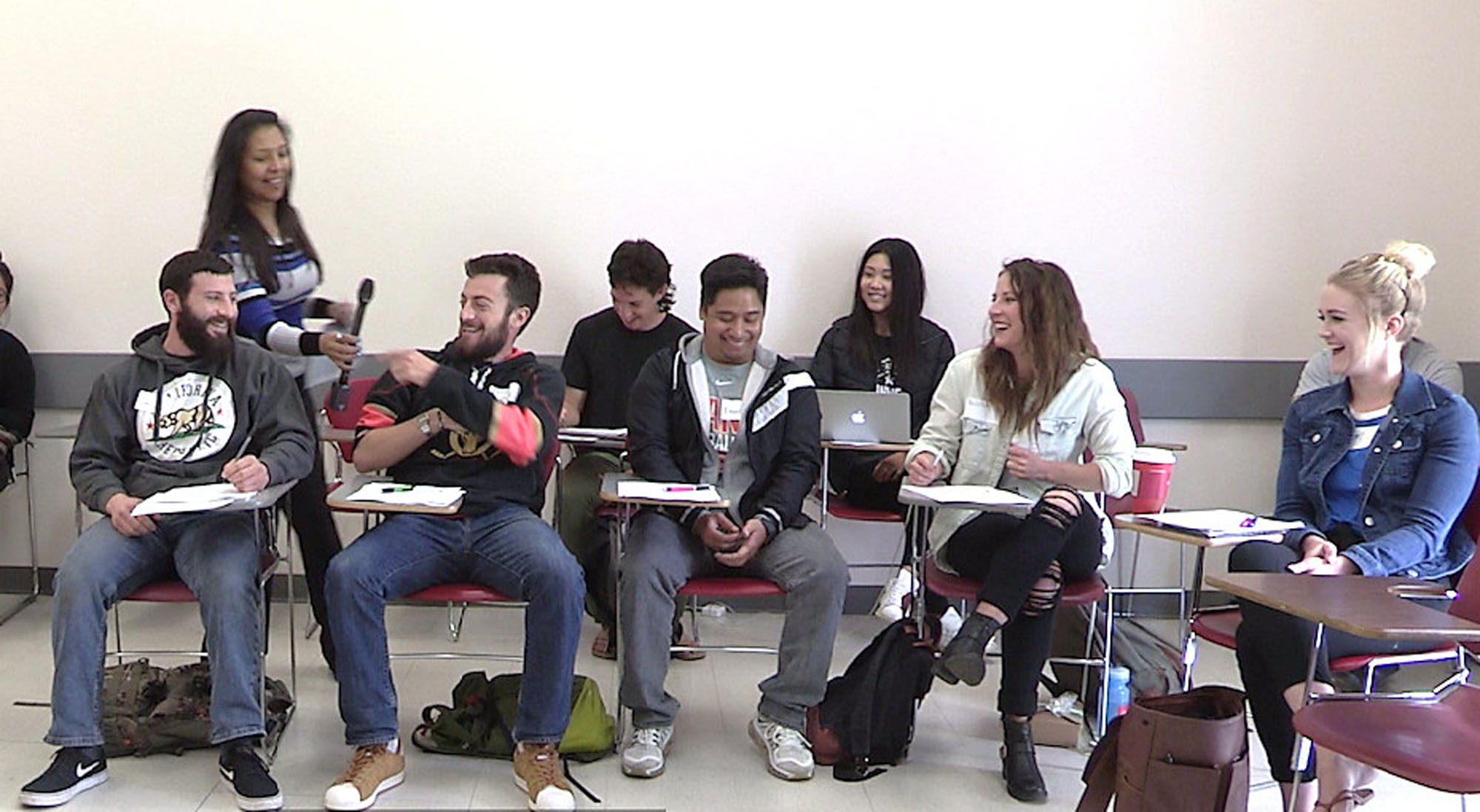 A group of smiling and laughing people in a classroom handing a microphone to each other to speak