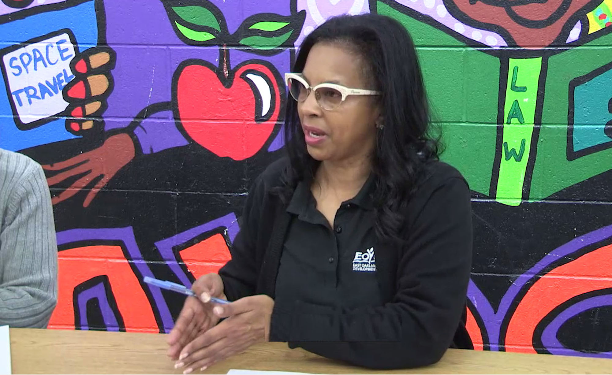 A woman speaking in a classroom with a mural painted on the wall behind her