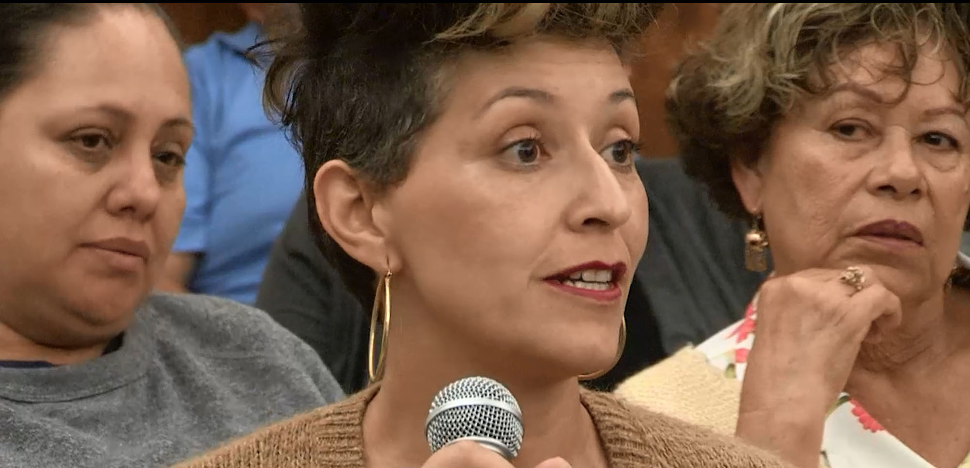 A woman holding and speaking into a microphone with two women sitting behind her