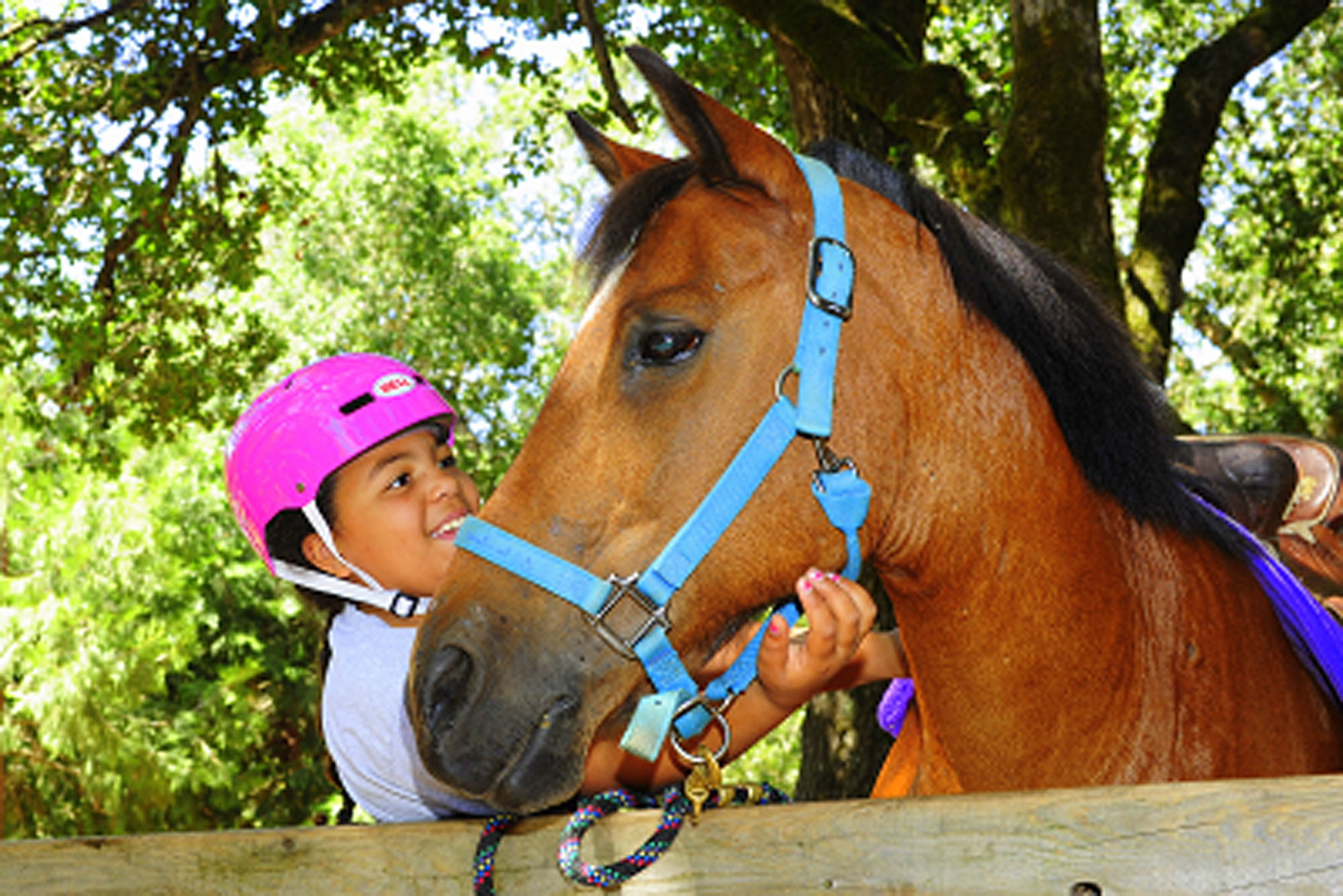 A child wearing a pink helmet smiles as they pet a horse