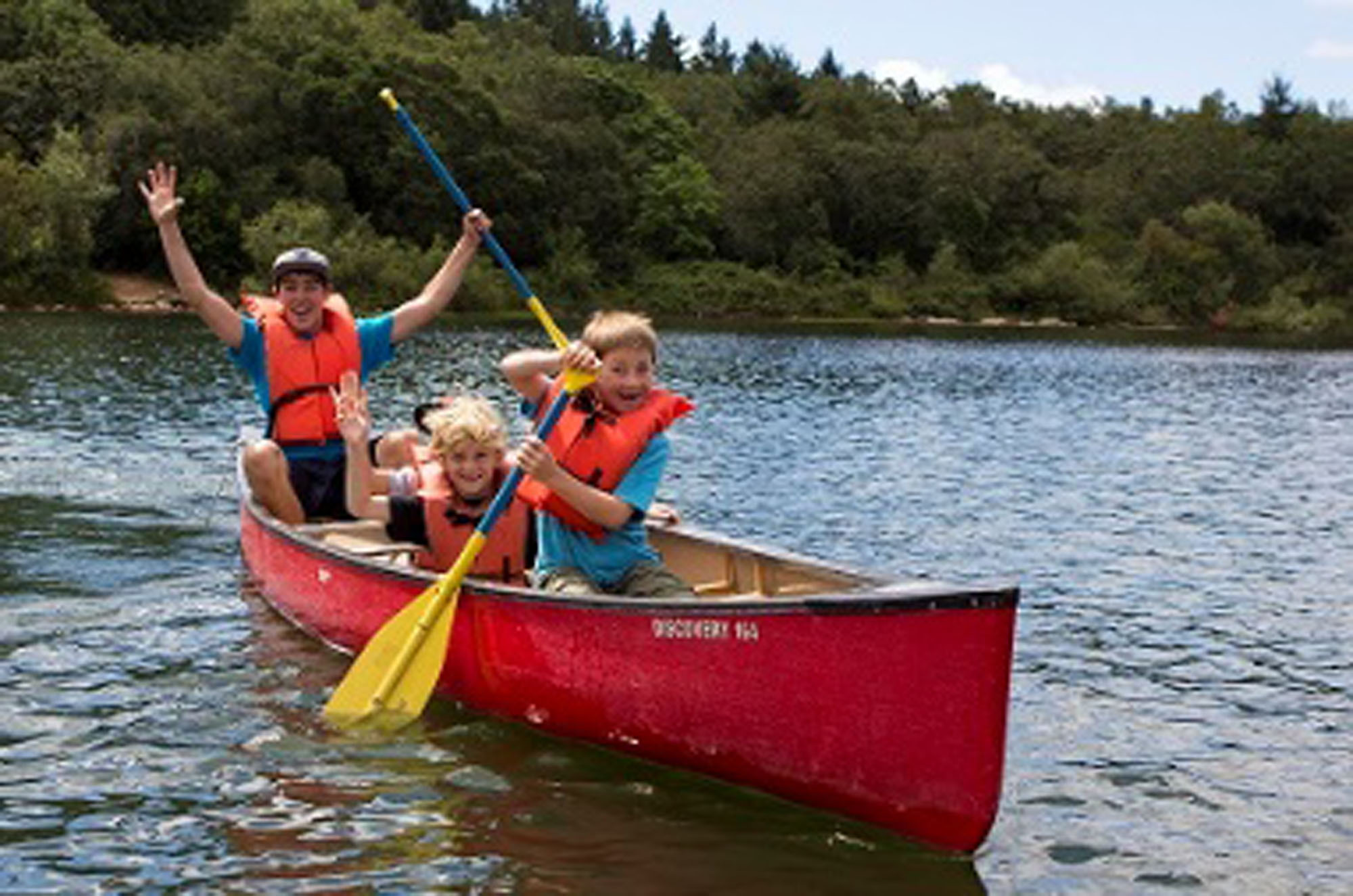 A teenager and a few children paddle a red canoe on a lake with a forest in the background